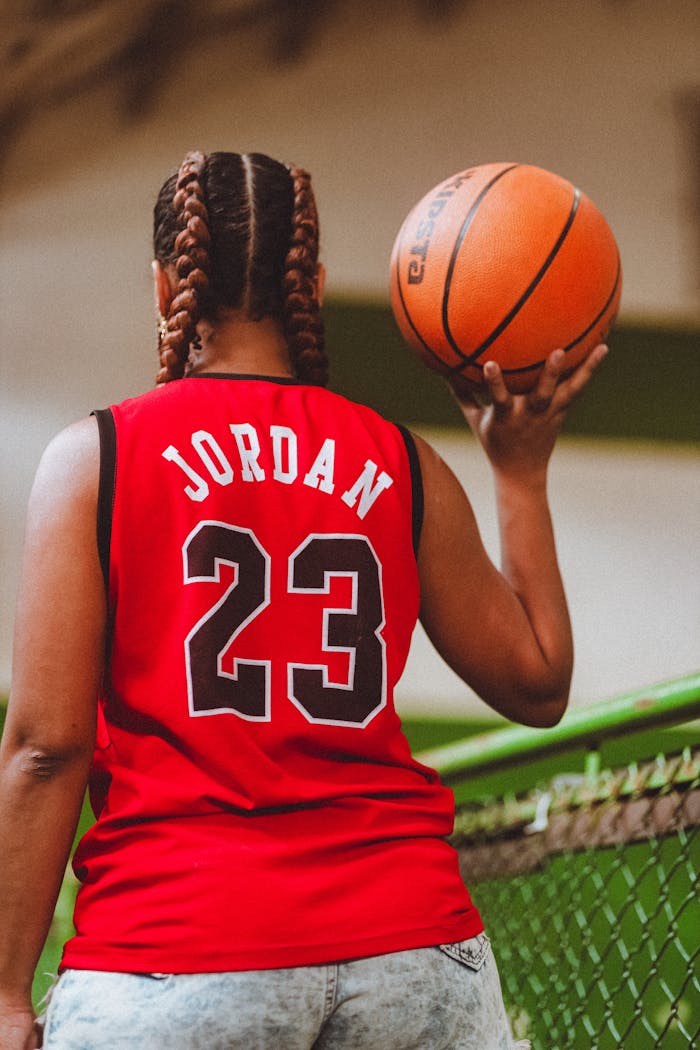 Services Woman in red sports jersey holding a basketball indoors, seen from behind.