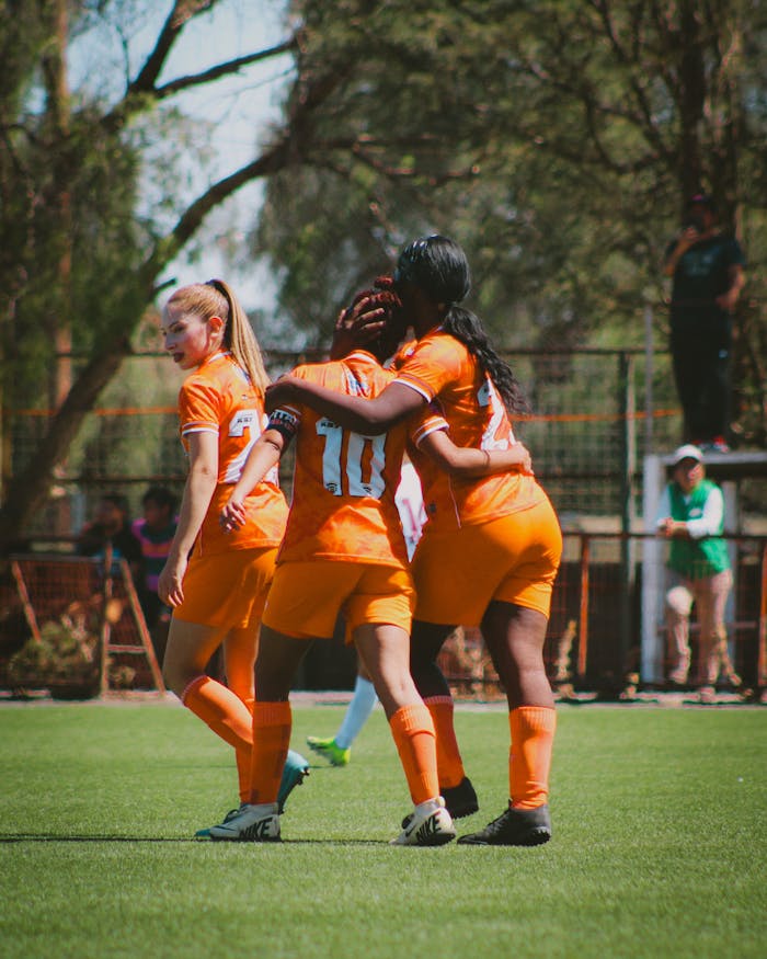 Services A women's soccer team in orange jerseys celebrates a goal during a match in the bright daytime.
